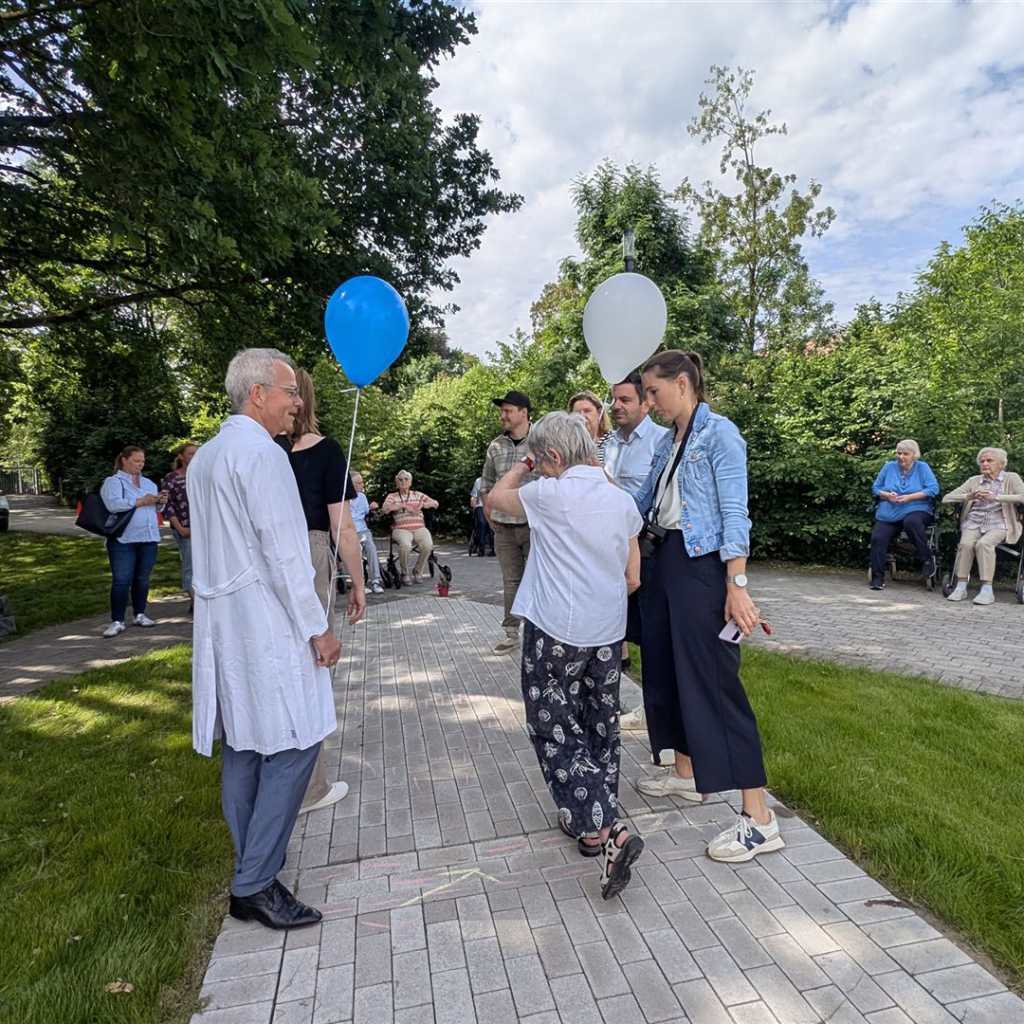 Menschen stehen draußen, einige mit Ballons, und ältere Menschen sitzen in Rollstühlen.