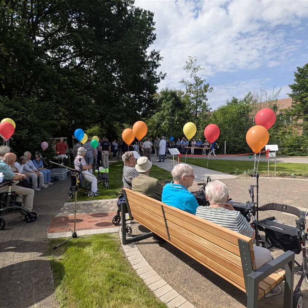 Eine Gruppe von älteren Menschen sitzt im Freien, einige auf einer Bank, andere in Rollstühlen oder mit Gehhilfen, umgeben von bunten Ballons und Bäum