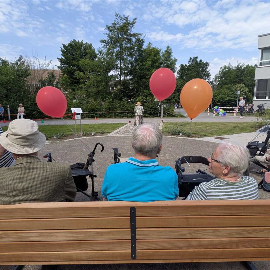 Ältere Menschen sitzen auf Bänken im Freien, umgeben von Bäumen und einem Gebäude, mit bunten Ballons in der Luft.