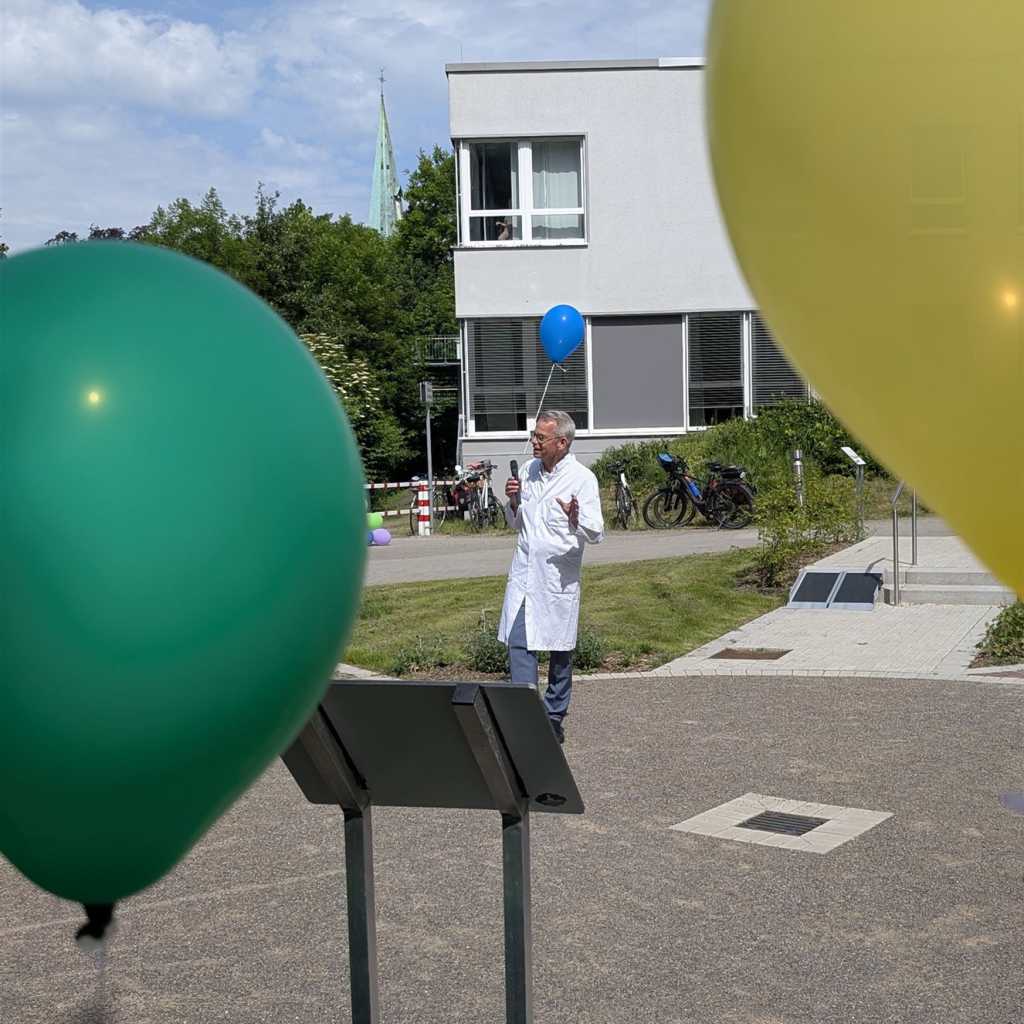 Mann in einem weißen Kittel hält einen blauen Luftballon und spricht in ein Mikrofon, während im Vordergrund grüne und gelbe Luftballons zu sehen sind