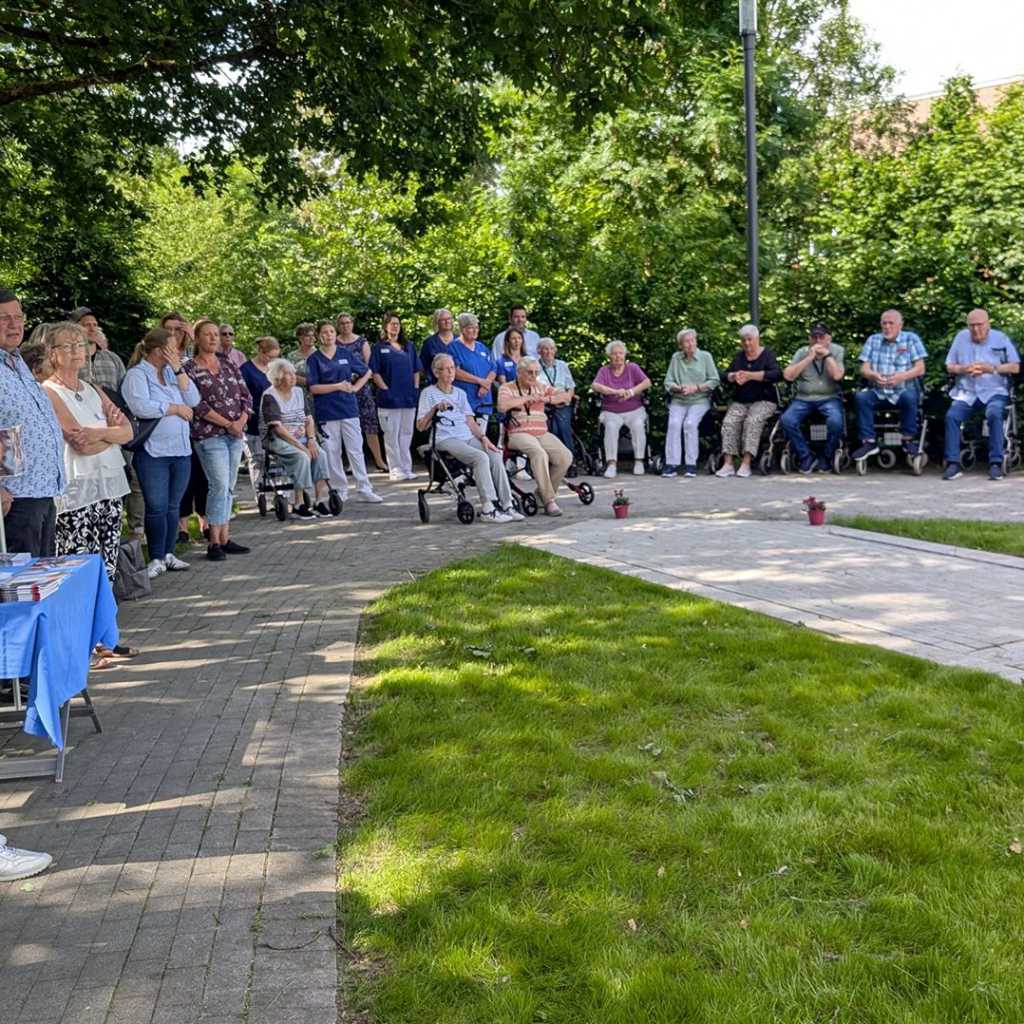 Eine Gruppe älterer Menschen sitzt im Freien, einige in Rollstühlen, während andere stehen und zuhören.