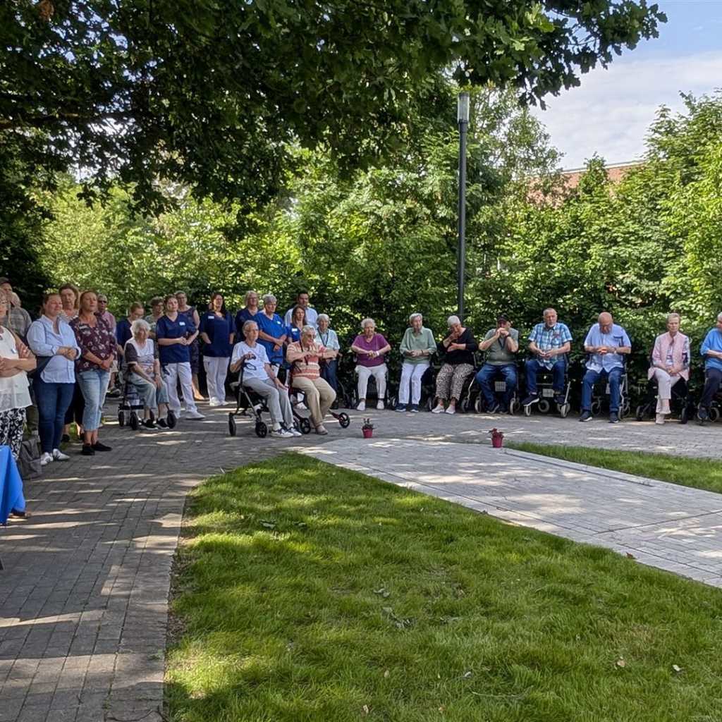 Eine Gruppe älterer Menschen sitzt in Rollstühlen und auf Stühlen im Freien, während eine jüngere Gruppe von Pflegekräften oder Freiwilligen daneben s