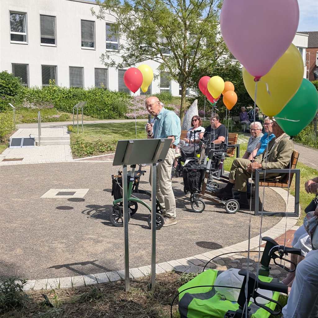 Ein älterer Mann spricht in ein Mikrofon, während er hinter einem Pult steht, daneben ein Rollator. Im Hintergrund sitzen mehrere ältere Personen auf 
